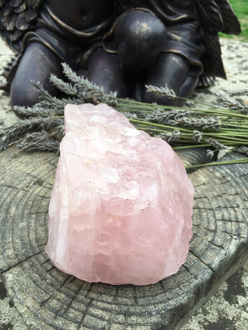 May include: A large, rough-hewn rose quartz crystal, displaying a soft pink hue. The crystal sits on a weathered, gray wooden surface, with dried lavender sprigs and a bronze statue in the background. The stone's texture is visible.