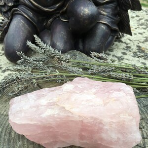 May include: A large, rough-cut rose quartz crystal in shades of pink rests on a weathered wooden surface. Dried lavender sprigs are arranged nearby, with a bronze statue of cherubs in the background. The crystal's texture is visible.