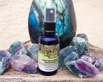 Puede incluir: Una botella de vidrio azul con una boquilla de pulverización, etiquetada "Crystal Cleansing/Charging Herbal Energy Spray" por Lorraine Tricksey. La botella está rodeada de cristales de fluorita verdes y morados.