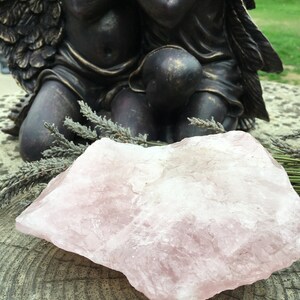 May include: A large, rough-hewn rose quartz crystal rests on a weathered wooden surface, with sprigs of dried lavender nearby. A bronze-colored statue of two cherubs with wings is in the background. The crystal is a soft pink color.
