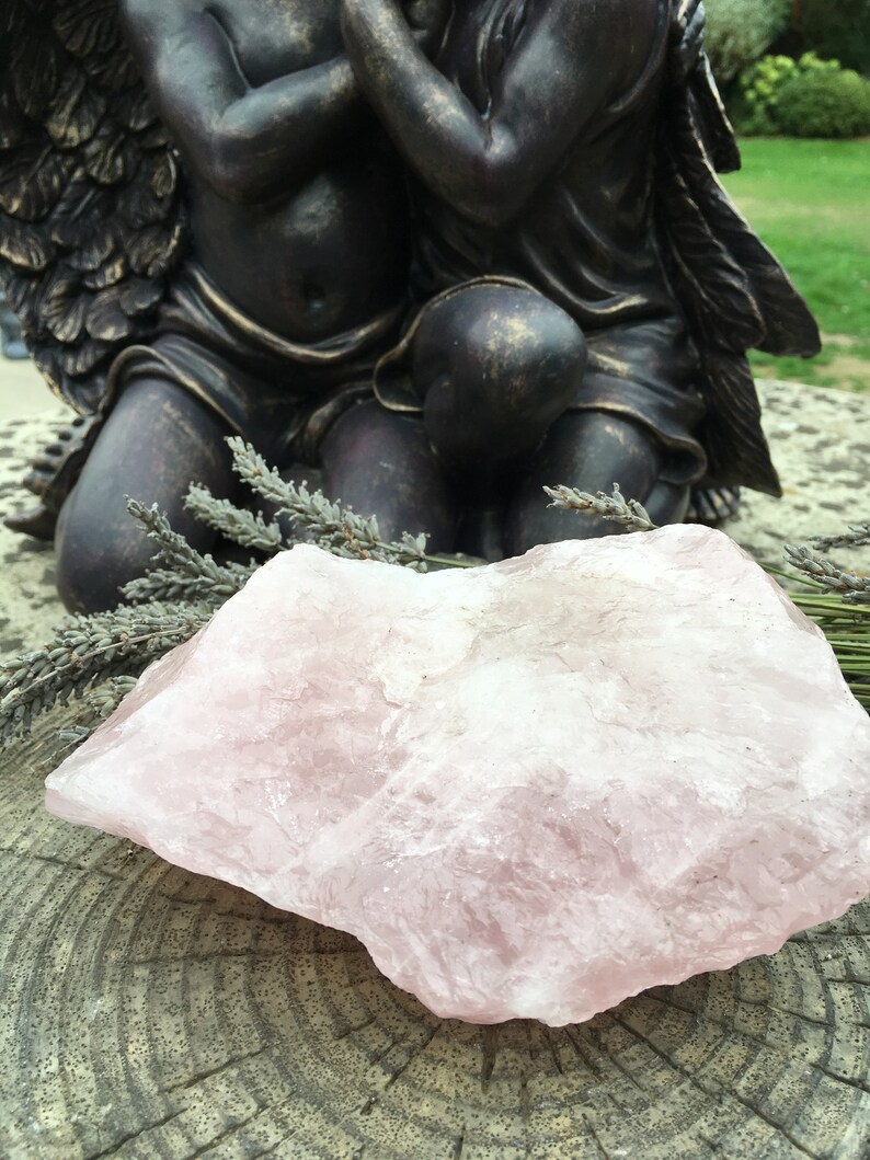 May include: A large, rough-hewn rose quartz crystal rests on a weathered wooden surface, with sprigs of dried lavender nearby. A bronze-colored statue of two cherubs with wings is in the background. The crystal is a soft pink color.