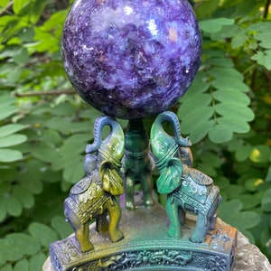 May include: A large purple amethyst sphere sits on a decorative stand made of three elephants. The elephants are painted in shades of green, blue, and gold.
