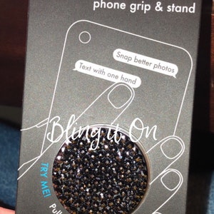 May include: Black PopSocket phone grip and stand with a sparkly, blinged-out design. The packaging features the text "Snap better photos" and "Text with one hand". The PopSocket also says "Try Me! Pull twice to expand".
