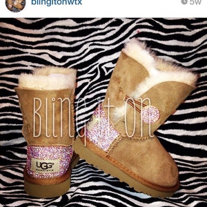 May include: A pair of brown UGG boots with faux fur trim. The boots are decorated with pink and white rhinestones. The boots are sitting on a black and white zebra print background. The text "UGG Australia" is visible on the boots.