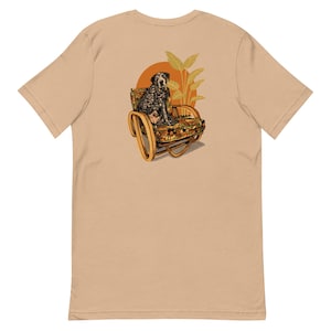May include: A tan t-shirt featuring a graphic of a dog sitting in a patterned chair with a sunset and tropical leaves in the background. The dog has black spots and is facing forward. The chair has a wooden frame.