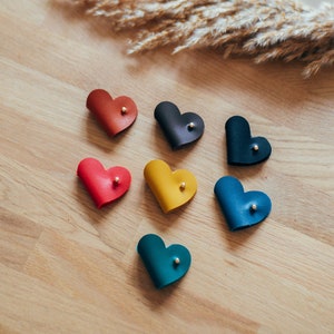 May include: Seven leather heart-shaped keychains in different colors: red, orange, yellow, green, blue, dark blue, and black. Each keychain has a small gold accent.