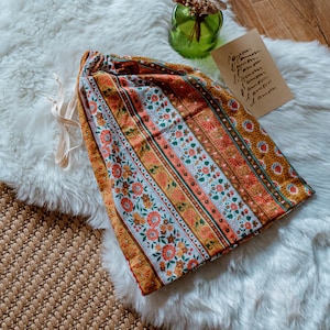 Peut inclure: Un sac à cordon avec un motif floral coloré à rayures orange, jaune et blanc. Le sac est en tissu et possède un cordon de serrage crème. Le sac est posé sur un tapis blanc et duveteux.