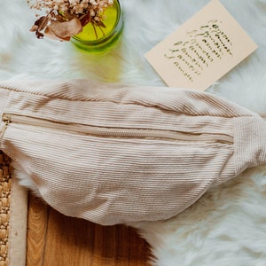 Puede incluir: Una riñonera de pana beige con cierre de cremallera. La riñonera está sobre una superficie blanca y esponjosa.