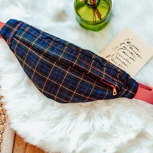 May include: A red and blue plaid fanny pack with a zippered pocket and a red strap. The fanny pack is lying on a white fluffy surface.