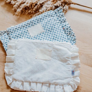 May include: Two white fabric pouches with ruffled edges. The top pouch has a blue and white geometric pattern and the bottom pouch has a plain white surface. Both pouches have a white label sewn on the front.