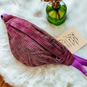 Puede incluir: Una riñonera de pana morada con una correa morada y una hebilla plateada. La riñonera está sobre una superficie blanca y esponjosa.