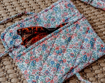 May include: A white quilted fabric pouch with a floral pattern and a brown pair of sunglasses inside. The pouch has a drawstring closure and a long strap.