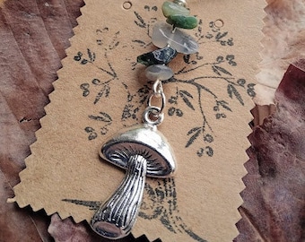 May include: A silver necklace with a mushroom charm and green and gray gemstone beads. The necklace is displayed on a brown paper tag with a tree design.