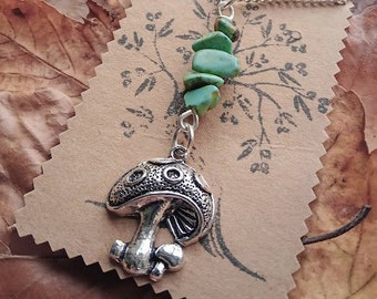 May include: A silver necklace with a mushroom charm and green gemstone beads. The necklace is displayed on a brown paper card with a tree design.