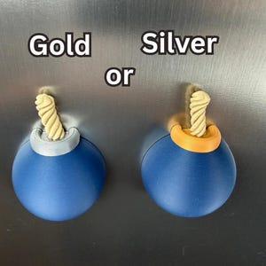 May include: Two spherical blue magnets shaped like cartoon bombs with a beige twisted fuse protruding from the top. One bomb features a silver-coloured top accent, and the other features a gold-coloured top accent. The text Gold or Silver is visible.