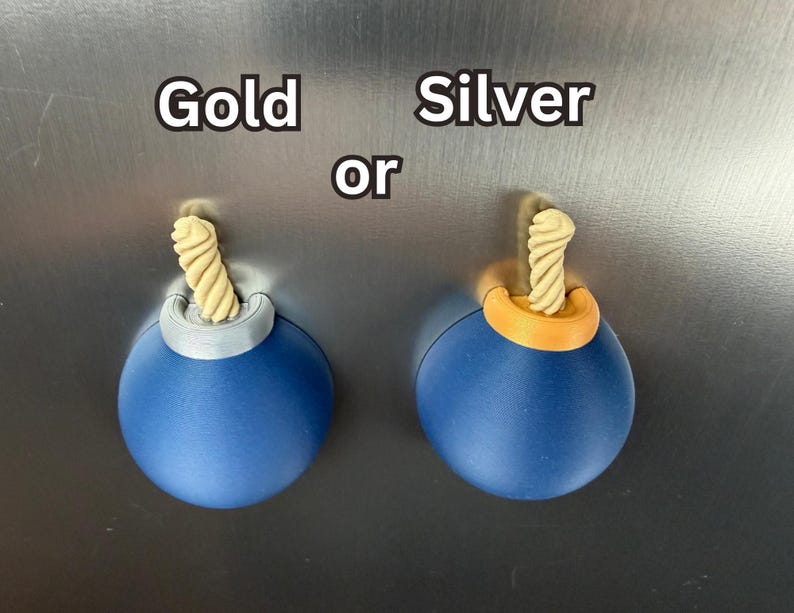 May include: Two spherical blue magnets shaped like cartoon bombs with a beige twisted fuse protruding from the top. One bomb features a silver-coloured top accent, and the other features a gold-coloured top accent. The text Gold or Silver is visible.