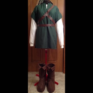 May include: A green tunic with brown leather straps and a white long-sleeved shirt underneath. The tunic is on a red stand with brown boots on the floor.