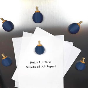 May include: Five dark blue bomb-shaped magnets with gold-coloured tops and tan rope fuses. One magnet secures a stack of white paper printed with the black text: Holds Up to 3 Sheets of A4 Paper! The other four magnets are arranged around the paper.
