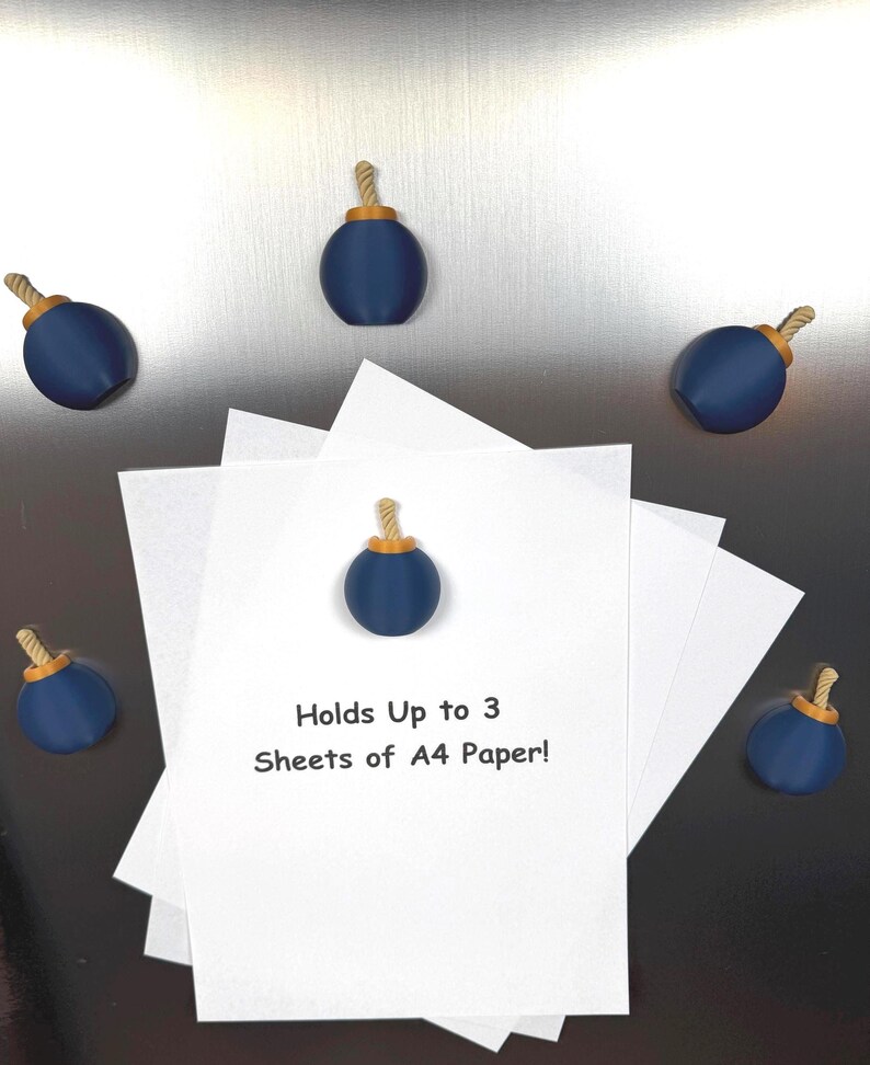 May include: Five dark blue bomb-shaped magnets with gold-coloured tops and tan rope fuses. One magnet secures a stack of white paper printed with the black text: Holds Up to 3 Sheets of A4 Paper! The other four magnets are arranged around the paper.