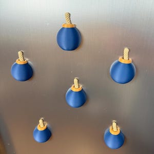 May include: A set of five spherical magnets shaped like cartoon bombs. Each magnet features a smooth dark blue body, a small orange circular top, and a short, textured beige rope fuse protruding from the centre of the orange top piece.