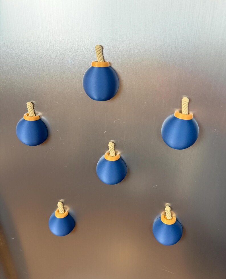 May include: A set of five spherical magnets shaped like cartoon bombs. Each magnet features a smooth dark blue body, a small orange circular top, and a short, textured beige rope fuse protruding from the centre of the orange top piece.