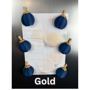 May include: Six spherical navy blue magnets with gold-coloured rope-textured tops arranged on a rectangular shopping list notepad. The notepad features a light yellow citrus fruit pattern and the word shop in cursive script. The text Gold is at the bottom.