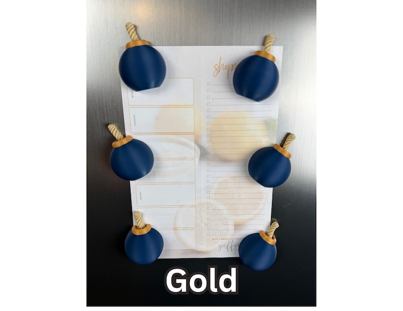 May include: Six spherical navy blue magnets with gold-coloured rope-textured tops arranged on a rectangular shopping list notepad. The notepad features a light yellow citrus fruit pattern and the word shop in cursive script. The text Gold is at the bottom.