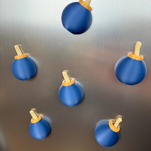 May include: A set of five spherical magnets shaped like cartoon bombs. Each magnet features a smooth, matte blue body, a bright orange circular base at the top, and a short, textured, light beige fuse protruding from the centre of the orange base.