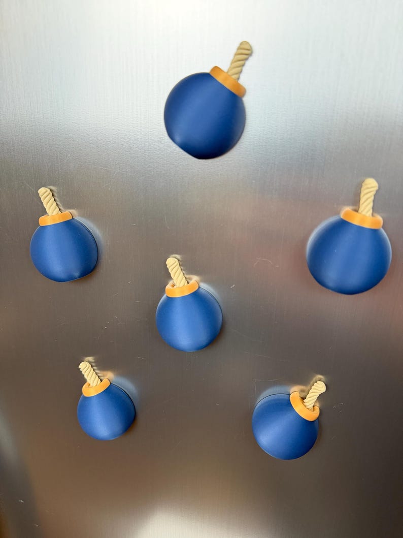 May include: A set of five spherical magnets shaped like cartoon bombs. Each magnet features a smooth, matte blue body, a bright orange circular base at the top, and a short, textured, light beige fuse protruding from the centre of the orange base.