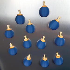 May include: A collection of eleven spherical magnets shaped like cartoon bombs. Each magnet features a smooth, matte blue body, a small orange circular base at the top, and a short, twisted beige fuse protruding from the centre of the orange base.