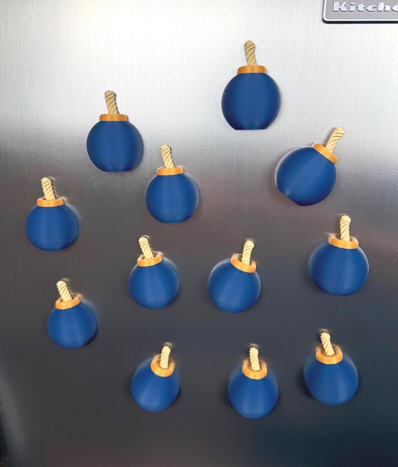 May include: A collection of eleven spherical magnets shaped like cartoon bombs. Each magnet features a smooth, matte blue body, a small orange circular base at the top, and a short, twisted beige fuse protruding from the centre of the orange base.