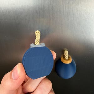 May include: Two spherical bomb-shaped magnets with a dark blue body and a light tan twisted fuse. One magnet is held in the foreground, featuring a grey top piece, while the other is attached to a metallic surface with a gold-coloured top piece.
