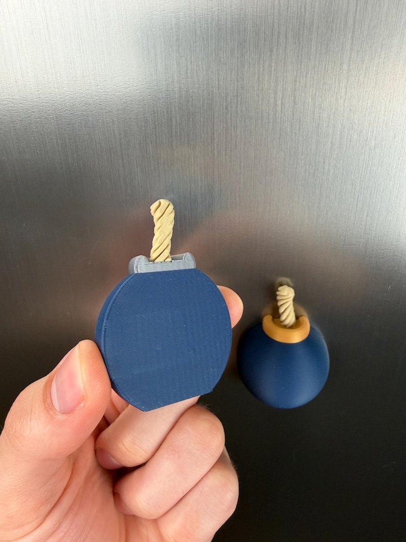 May include: Two spherical bomb-shaped magnets with a dark blue body and a light tan twisted fuse. One magnet is held in the foreground, featuring a grey top piece, while the other is attached to a metallic surface with a gold-coloured top piece.