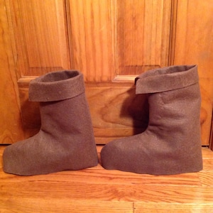 May include: A pair of brown felt boots with a folded cuff. The boots are sitting on a wooden floor.
