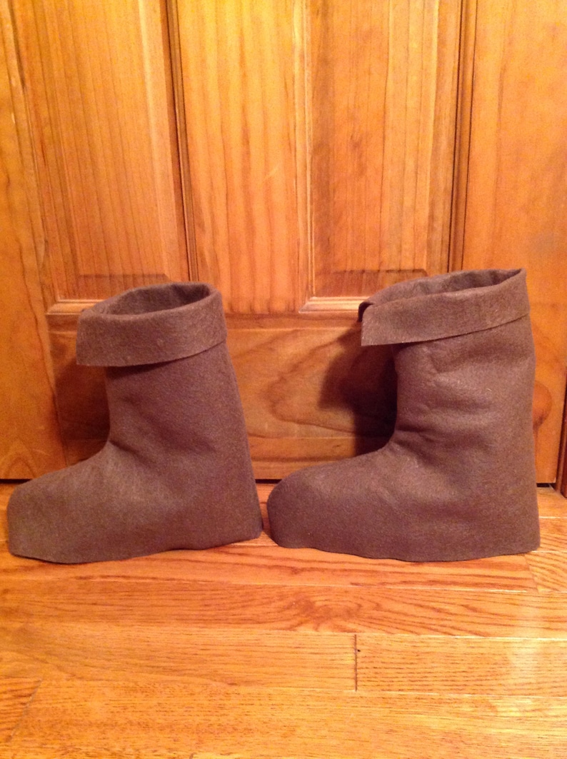 May include: A pair of brown felt boots with a folded cuff. The boots are sitting on a wooden floor.