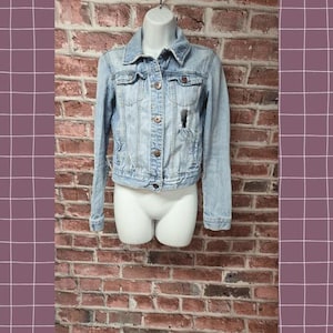 May include: Light blue denim jacket with a classic collar, button closure, and distressed details. The jacket is displayed on a white mannequin against a brick wall background.