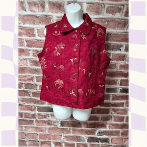 May include: A red sleeveless vest with embroidered designs of various shapes and objects. The vest has a collared neckline and button closures down the front. The embroidery is in a light tan color.