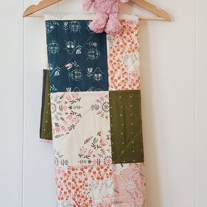 May include: A patchwork baby quilt with a variety of floral and geometric patterns in shades of green, pink, white, and orange. The quilt is hanging on a wooden hanger with a stuffed pig toy.