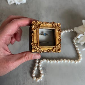 May include: A gold ring with a rectangular blue gemstone is displayed inside a small, ornate, gold-framed box. The box is held by a hand, with a pearl necklace and white flowers in the background.