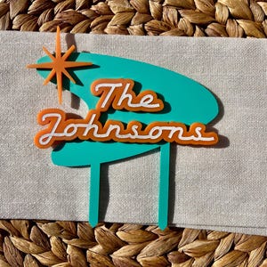 May include: A teal and orange cake topper with the text "The Johnsons" in a retro font. The topper features a starburst design and is made of acrylic. The topper is designed to be inserted into a cake.