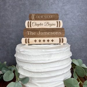 May include: A white frosted cake topped with a wooden book-shaped cake topper. The topper has four stacked books with the phrases "Our Next", "Chapter Begins", "The Jamesons", and a decorative book with diamonds.