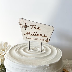 May include: A wooden cake topper with a retro sign design. The sign reads "The Millers" and "October 12th, 2025".