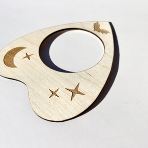 Puede incluir: Una planchette de madera con una luna creciente, estrellas y un dise&ntilde;o de murci&eacute;lago. La planchette se utiliza para comunicarse con los esp&iacute;ritus durante una sesi&oacute;n de espiritismo.