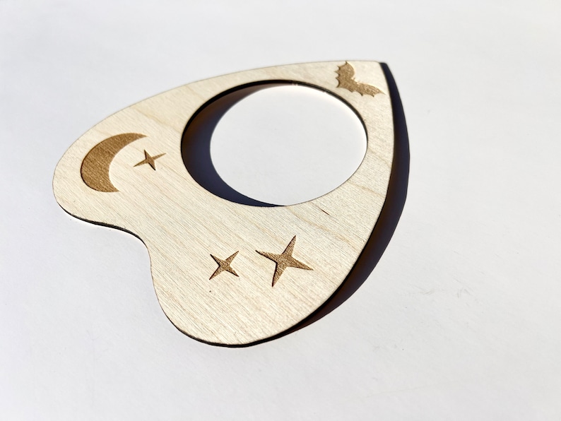 Puede incluir: Una planchette de madera con una luna creciente, estrellas y un dise&ntilde;o de murci&eacute;lago. La planchette se utiliza para comunicarse con los esp&iacute;ritus durante una sesi&oacute;n de espiritismo.