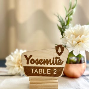 May include: Wooden table number sign with the word "Yosemite" in a retro font, and the text "TABLE 2". The sign has a dark brown border and a National Park Service emblem. The sign is on a wooden base.