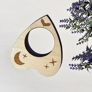 Puede incluir: Una planchette de madera con un dise&ntilde;o de luna creciente, estrellas y un murci&eacute;lago. La planchette se utiliza para comunicarse con los esp&iacute;ritus durante una sesi&oacute;n de tablero Ouija.