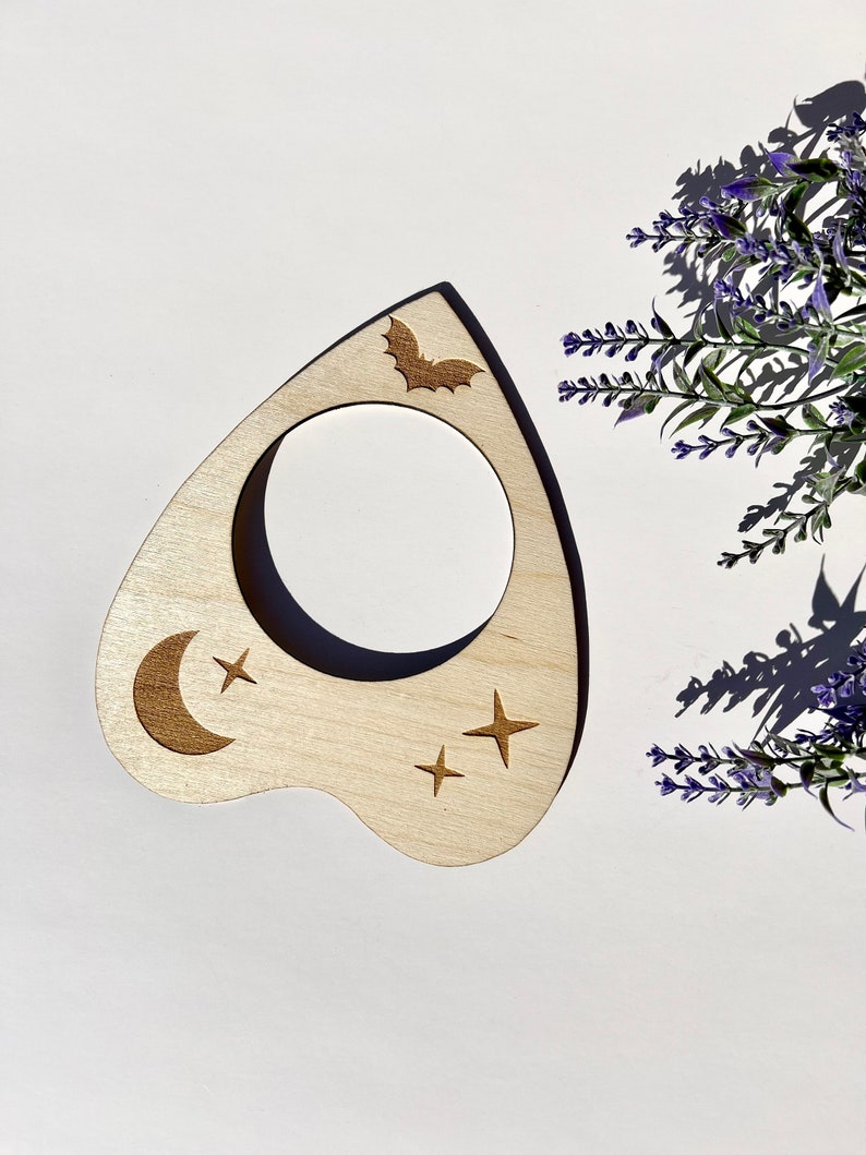 Puede incluir: Una planchette de madera con un dise&ntilde;o de luna creciente, estrellas y un murci&eacute;lago. La planchette se utiliza para comunicarse con los esp&iacute;ritus durante una sesi&oacute;n de tablero Ouija.