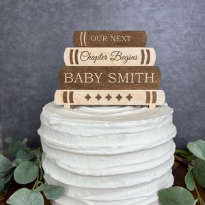 May include: A white frosted cake topped with a wooden cake topper. The topper is designed to look like a stack of books. The books are engraved with the words "OUR NEXT", "Chapter Begins", and "BABY SMITH".