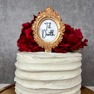 May include: A white frosted cake with a gold frame cake topper that reads "Til Death". The cake is decorated with red flowers. The cake is on a wooden cake stand.