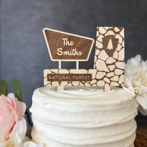 May include: A wooden cake topper with a sign that reads "The Smiths" and "National Forest." The topper has a rustic design with a stone-like texture and a tree emblem. The cake is white with a ruffled edge.